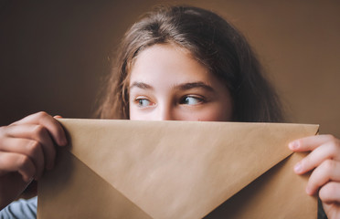 A young girl looks out from behind the envelope. The girl received the envelope and is happy.