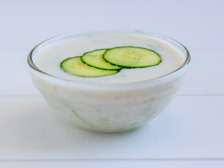 Three slices of cucumber in a bowl with tzatziki sauce on white wooden table.