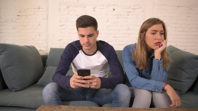 panning shot of young man using social media on mobile phone ignoring his girlfriend at home sofa couch with the woman frustrated upset bored feeling ignored by boyfriend in internet addiction concept