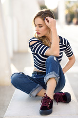 Blonde woman, model of fashion, sitting in urban background.