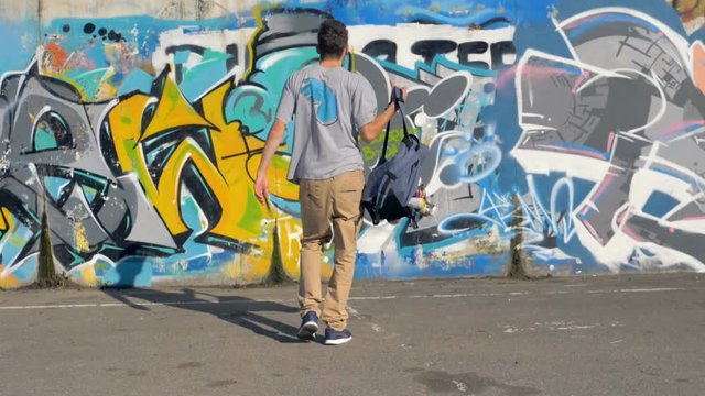 Graffiti artist is coming up to the  wall covered with graffiti pictures, with a bag full of spray paints.