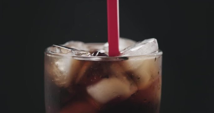 Slow motion closeup put red straw into cola with ice cubes in glass ove black background