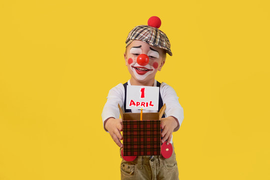 Kid clown with red nose 5 years on the bright yellow background. Little child holds box with white sheet, text 1 April. Concept birthday, holiday, celebration, party. Happy jester looking small box.