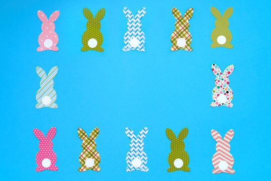 Easter bunny decoration on blue background, copy space. DIY holiday handicraft of colorful rabbits. Flat lay, top view. Border of paper rabbits cutouts. Easter greeting card. Happy Easter concept.