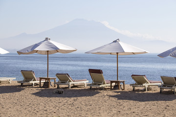 Tropical sand beach with sun loungers and umbrellas with a view of the volcano next to the sea water in island Bali, Indonesia