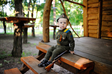 Child boy in military uniform sits on a wooden house and forest background. Victory Day, February 23, May 9, soldier