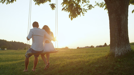 LENS FLARE: Man and woman enjoying view of a summer sunset on a romantic swing.