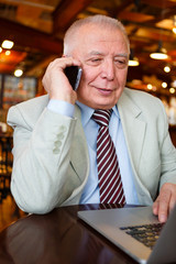 Old senior businessman in suit and tie, sitting in city cafe house talking by cell phone