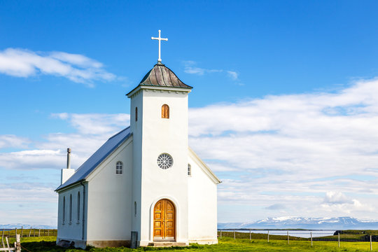 Flateyjarkirkja white lutheran church with meadow in foreground and sea  fjord with blue sky and mountains in the background, Flatey, Iceland