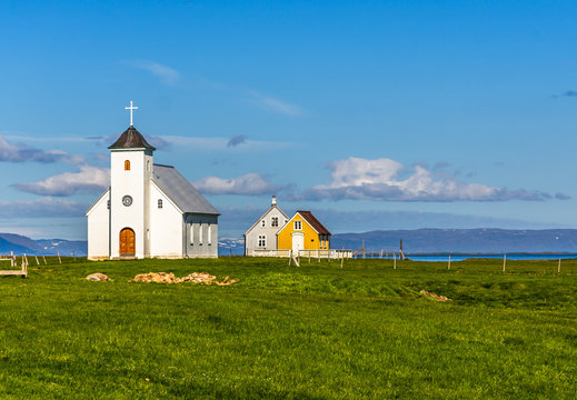 Flateyjarkirkja white lutheran church and couple of living huts with meadow in foreground and sea  fjord with blue sky in the background, Flatey, Iceland
