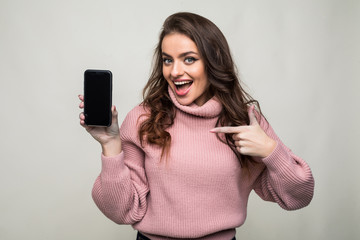 Portrait of casual girl pointing finger at blank screen mobile phone isolated over gray background