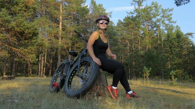 Fat bike also called fatbike or fat-tire bike in summer riding in the forest. Beautiful girl and her bicycle in the forest. She sits on the stump and rests from the trip.