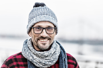 Portrait of urban man in winter knitted wear. Portrait of man in glasses and knitted white-blue scarf and hat. Portrait of happy man. Smiled non shaved guy on outdoor background