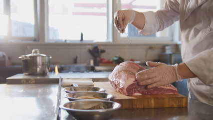 Butcher adding spices into large piece of fresh raw meat lying on a wooden board in a commercial kitchen