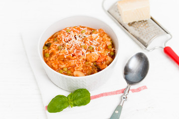 Bowl of Minestrone, a traditional Italian vegetable soup made of carrots, peas, beans, celery, tomatoes, garlic and onions with grated parmesan cheese and basil on white kitchen table