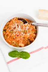 Bowl of Minestrone, a traditional Italian vegetable soup made of carrots, peas, beans, celery, tomatoes, garlic and onions with grated parmesan cheese and basil on white kitchen table