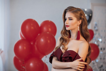 Portrait of an elegant woman in a gray room on a background of red and silver balloons.Attractive woman in an open, burgundy dress posing alone, standing in a set, beautiful room.Valentine's Day