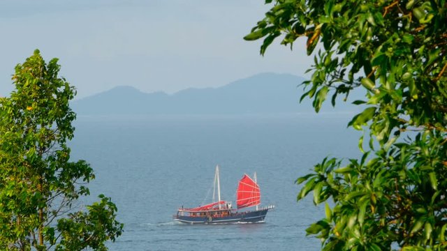 Traditional chinese ship with red sail floats along the sea at sunset or sunrise time. Island is visible on the horizon, green folliage on the foreground. Idyllic tropical exotic meditative landscape.