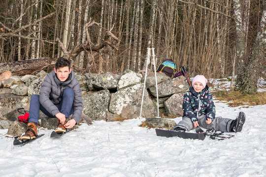 Winter activity. A boy and girl sitting in the snow putting on their skis against a stone wall.