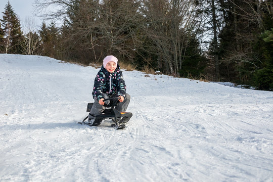 Winter snow activity. Front view of a girl riding a sledge downhill surrounded by trees.