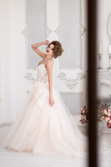 Fototapeta premium Morning of the bride. A beautiful bride is standing in a lace wedding dress. Wedding hairstyle and makeup. Hand at the face. Spinning in a dress. Photo through the window.