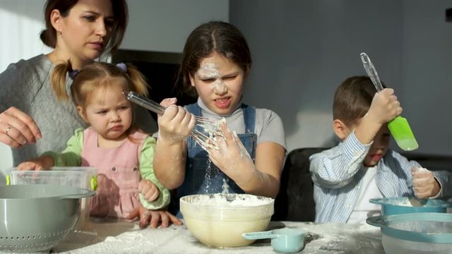 A beautiful mother with three cheerful children of different ages kneads the dough. The elder daughter removes the dough from the corolla