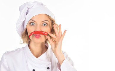 Spicy food concept - woman chef making moustache out of red hot chili pepper. Professional cook in white suit holds red hot chili pepper as mustache while looking at camera with wide eyes. Copy space.