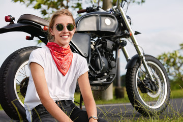 Fototapeta na wymiar Photo of cheerful female biker sits near black motorcycle in open air, wears stylish clothes, travels in unknown countryside place against wonderful scene background. Outdoor lifestyle concept.