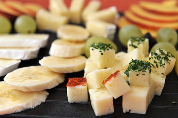 Fruit and cheese platter - black plate on a red table mat. Bananas, grapes, pear and peaches with brie and Gouda cheese.