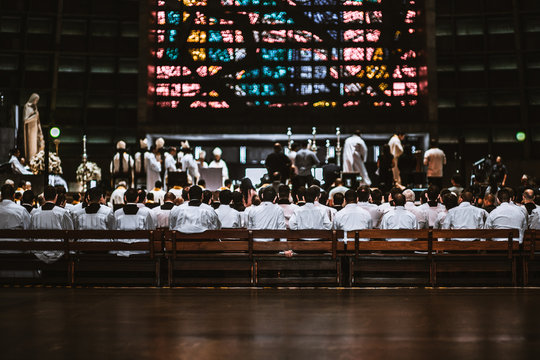 Rear view of the crowd of novices in white shirts sitting on the long wooden benches in a dark Catholic cathedral interior during a Christmas liturgy with a solemn ceremony in the defocused background