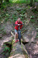Obraz premium young man in red jacket hiking in forest, climbing up to the log with bicycle
