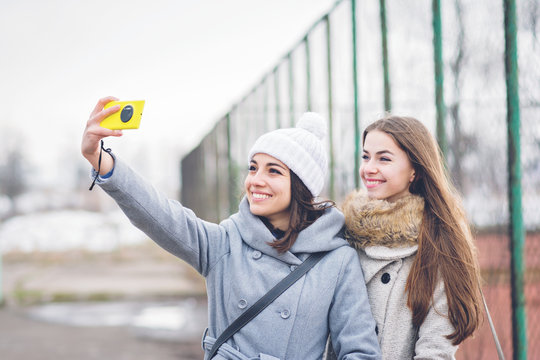 Two millennial teenage girls taking a selfie outdoors in winter using smart phone, smiling. No makeup, natural lighting