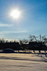 A field covered with snow with traces of paws, next to snow-covered trees and houses on a sunny winter day against a cloudy sky, Belarus