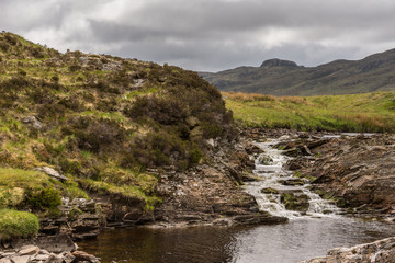Braemore, Scotland - June 8, 2012: Dundonnell Rvier rapids along A832 road offers bronwn rocks and grassy hills under dark cloudscape. Hills in back. Brown pool in front.