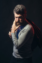 Elegant confident fashion man with beard and mustache in stylish gray pullover holds black leather jacket on shoulder. Pensive man with stylish hairdo stroking his beard. Isolated on black background.
