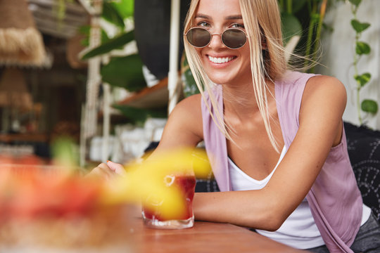 Indoor shot of happy blonde woman wears shades, sits against cozy cafe interior with cocktail, being satisfied with summer rest in tropical country, tastes exotic beverages. Pretty woman recreats