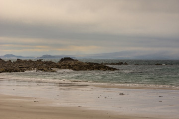 Mellon Udrigle, Scotland - June 8, 2012: Sandy Atlantic Ocean beach under thick brown-blue sky. Rocks in surf. Dark foggy hills in back.