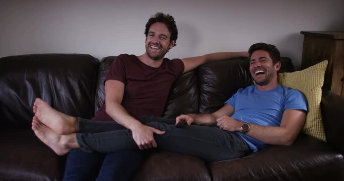 4K Gay male couple relaxing at home at night, watching tv & laughing. Slow motion.