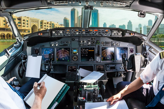 Airplane cockpit flying on Dubai skyscrapers skyline at sunset, United Arab Emirates, with pilots arms and blank white papers for copy space.
