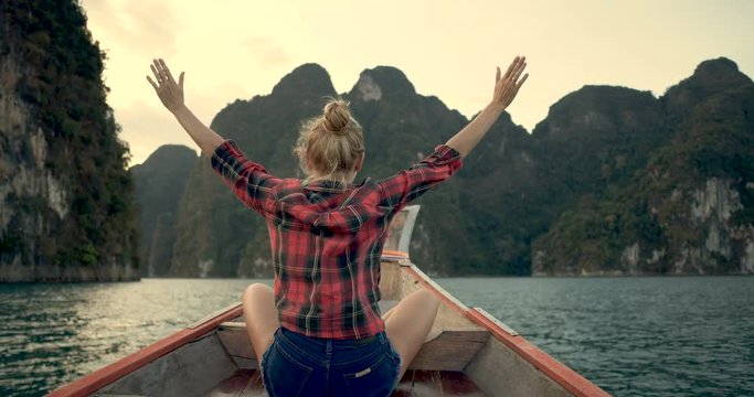 Back view of woman sitting in wood long tail boat with raised hands over tropical limestone cliffs and sunset background - video in slow motion
