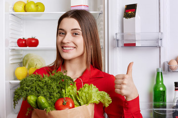 Photo of glad female shows ok sign and demonstrates approval, holds vegetables, comes from grocer`s...