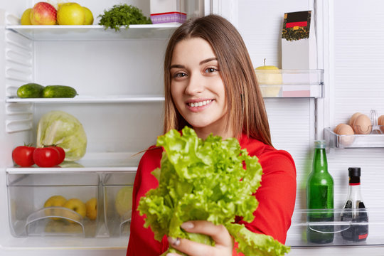 Positive young attractive woman holds lettuce, being vegeterian, stands against opened refrigerator full of fruit and vegetables, supports healthy eatting. Lovely female stands in kitchen near fridge