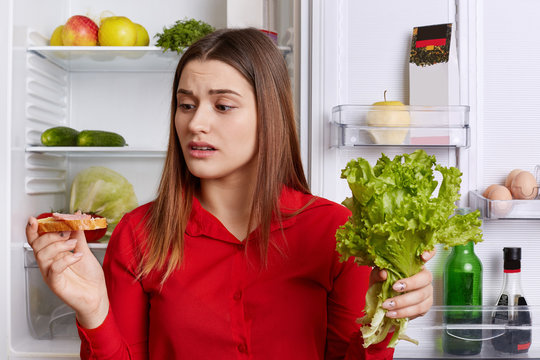 Horizontal shot of brunette female with upset expression looks at sandwhich, keeps lettuce, wants to eat meat, keeps to diet, doesn`t know what to eat for breakfast. Nutrition and people concet
