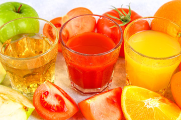 Glasses with fresh orange, apple, tomato juice on a gray concrete table. Lobules Fruits and vegetables around.