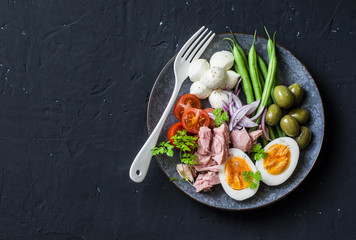 Balanced breakfast or snack - plate of canned tuna, green beans, mozzarella cheese, tomatoes, boiled egg, olives on a dark background, top view