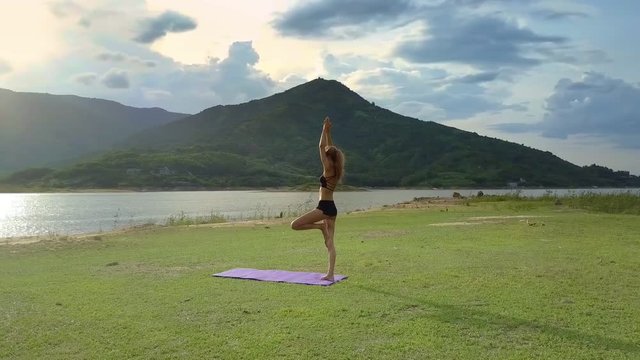 Flycam Shows Girl in Yoga Pose on Green Grass Lake Bank