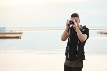 Stay where you are, this photo is amazing. Portrait of creative good-looking freelance photographer looking through camera while taking shots of nature and people, standing near seashore