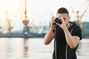 Attractive guy working with camera. Young stylish photographer looking through camera during photo session with gorgeous model, taking pictures in harbour near seashore, focusing on work