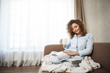 Woman decided to treat herself in bright good day. Portrait of attractive female curly-haired female sitting on sofa in pyjamas, drinking coffee, enjoying reading magazine, covering feet with blanket