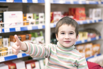 Happy boy 4 years in a supermarket on the background of shelves with chocolate. The boy showed that...